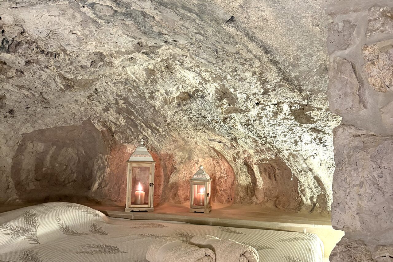La Grotta: a bed set into the raw rock niche, two lit lanterns on the ledge behind it and folded towels at the foot.