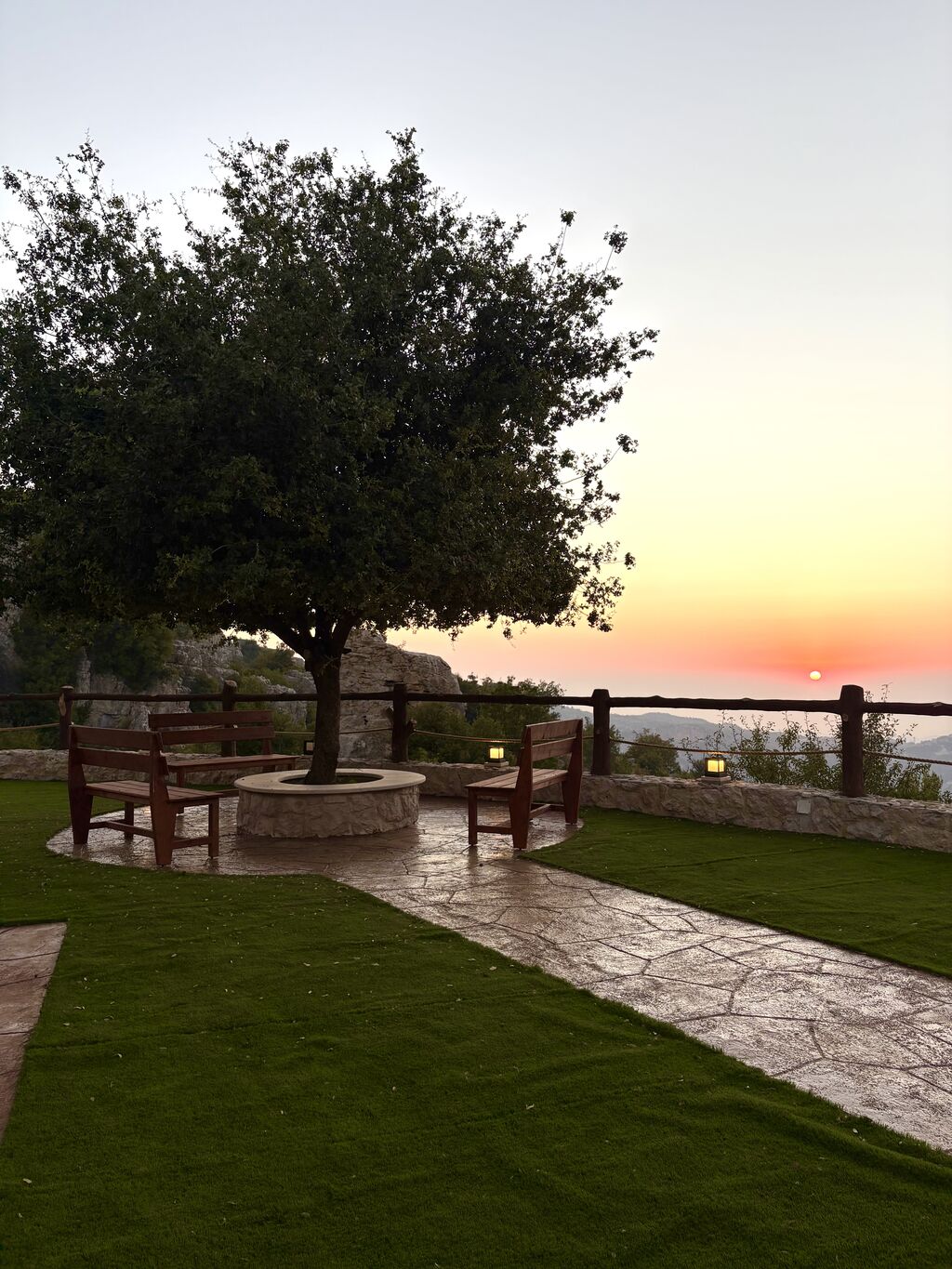 Wooden benches around an oak tree growing from a stone ring on the lawn at Cap de Douma, with a timber fence and the valley beyond at sunset.